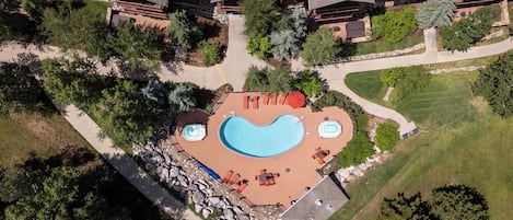 An aerial view of a resort with a large swimming pool, a hot tub, and outdoor seating areas surrounded by trees and a well-maintained lawn.