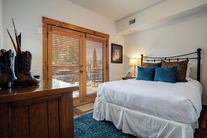 A bedroom with a bed, a wooden nightstand, a desk with boots, a window with blinds, and a picture on the wall. - EL5105 | Private Patio with Hot Tub | Air Chilling | Close to Skiing | 4 Pools (Steamboat Springs)