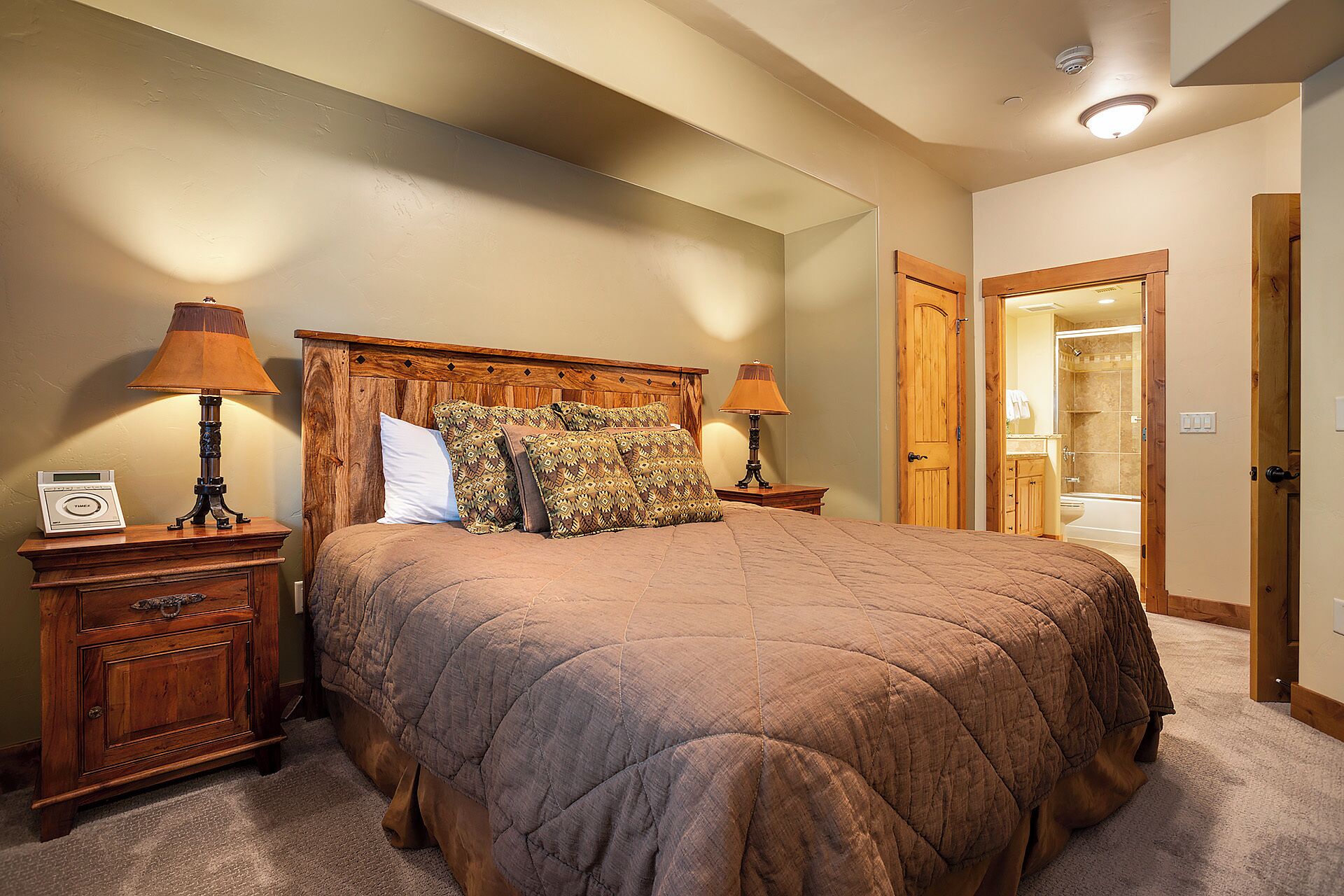 A bedroom with a large bed, two bedside tables with lamps, a wooden headboard, and a door leading to a bathroom.