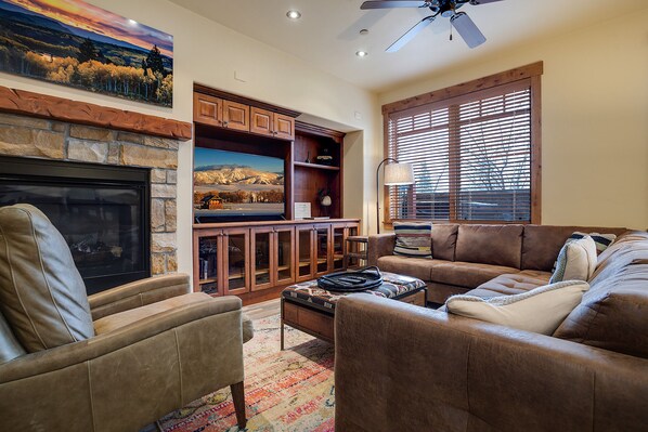 A living room with a fireplace, leather sofas, a television, and a window with blinds. - EL5111 | Private Courtyard with Hot Tub | Air Chilling | Close to Slope (Steamboat Springs)