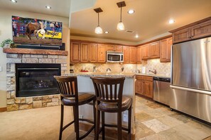 A kitchen with a stone fireplace, wooden cabinets, stainless steel appliances, and a bar area with two stools. - EL5114 | Close to Mtn Activities | Private Hot Tub | Air Chilling | 4 Pools (Steamboat Springs)