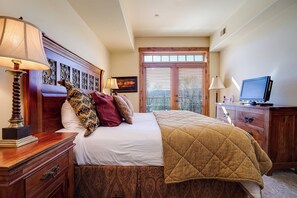 Habitación de hotel con cama, cabecera de madera, televisor, escritorio con lámpara y ventana con persianas.