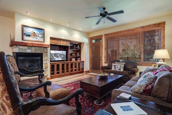 A cozy living room with a fireplace, a television, wooden furniture, and a ceiling fan. - EL5207 | Short Walk to Mtn| Air Chilling | 10 Hot Tubs| 4 Pools (Steamboat Springs)