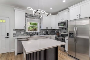 A modern kitchen with white cabinetry, a central island, stainless steel appliances, and a window with a view of greenery. - The Buford Luxe | Game Room + Office | 3BR (Atlanta)