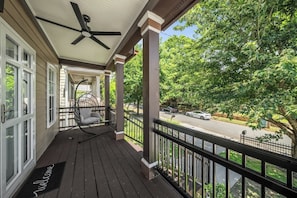 A covered porch with a ceiling fan, a rocking chair, and a view of a street with cars and trees. - The Buford Luxe | Game Room + Office | 3BR (Atlanta)