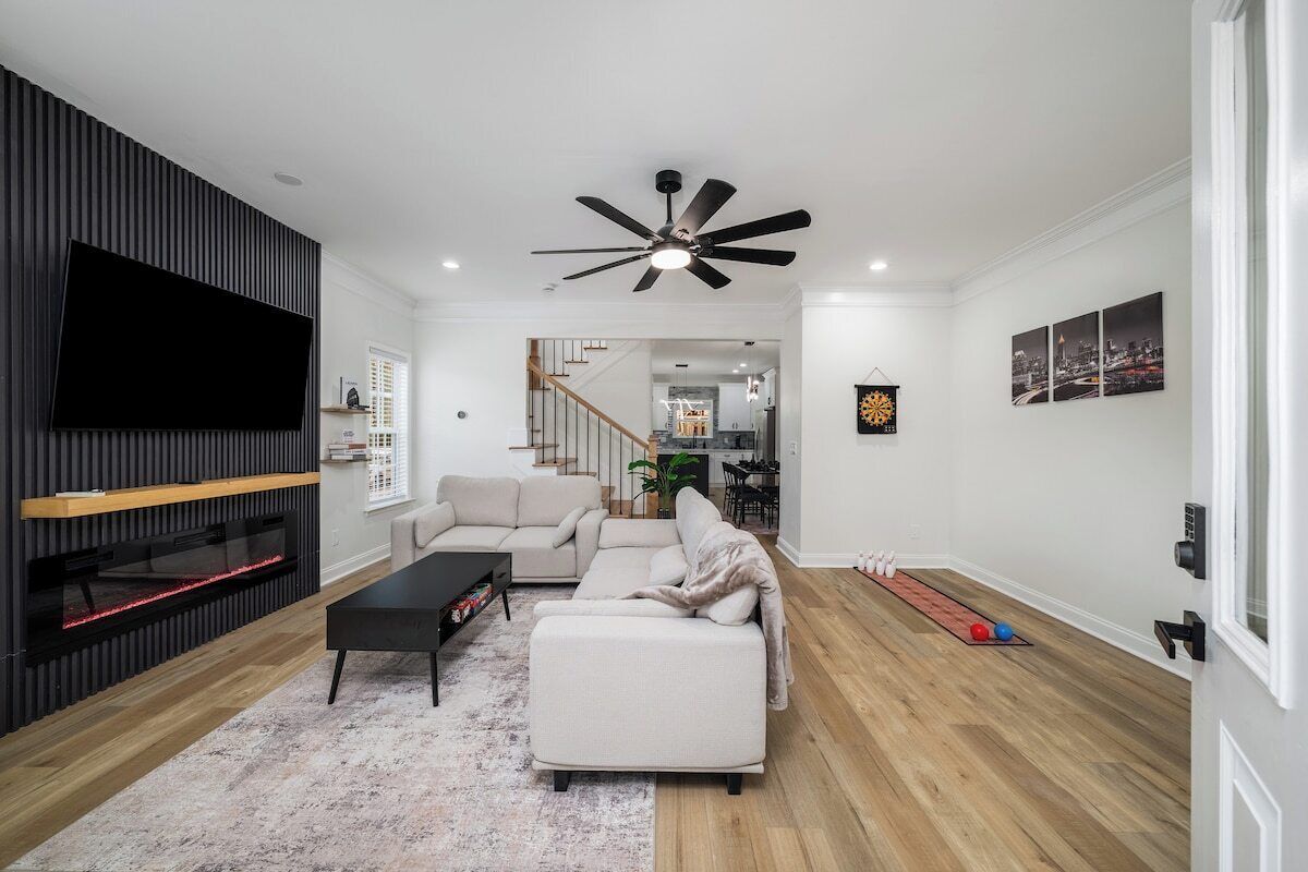 A modern living room with a flat-screen TV, a ceiling fan, a fireplace, and a dining area in the background.