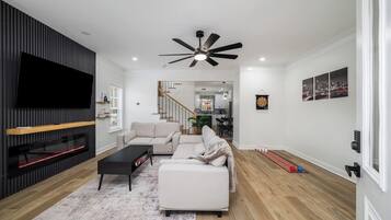 A modern living room with a flat-screen TV, a ceiling fan, a fireplace, and a dining area in the background.