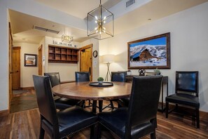 A dining area with a round wooden table, black leather chairs, a chandelier, a wall-mounted shelf, a painting of a snowy cabin, and a door labeled "UNLOAD HERE". - EL5208 | Spacious with Mountain View | Air Chilling | Close to Mtn | Hot Tubs (Steamboat Springs)