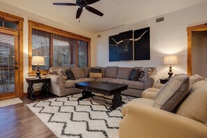 A living room with a sectional sofa, a coffee table, and two lamps. There are wooden blinds on the windows, and a ceiling fan is visible. - EL5209 | Gorgeous Second Floor Residence | Very Spacious | Air Chilling | Pool (Steamboat Springs)