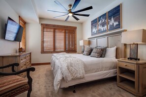 Ein Schlafzimmer mit Bett, Holz-Kopfteil, Nachttischen, Schreibtisch, Stuhl, Deckenventilator und an der Wand montiertem Fernseher.