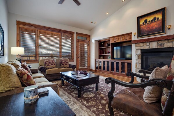 A spacious living room with a fireplace, wooden furniture, and a large window with blinds. - EL5303 | Mtn Getaway | Resort Amenities | 2 King Suites | Air Chilling (Steamboat Springs)