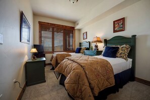 A bedroom with two beds, a nightstand, a lamp, a television, and a window with blinds. - EL5303 | Mtn Getaway | Resort Amenities | 2 King Suites | Air Chilling (Steamboat Springs)