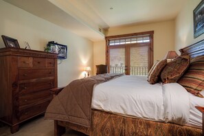 A bedroom with a wooden dresser, a bed with patterned bedding, a nightstand with a lamp, and a window with blinds. - EL5305 | Top Floor with Mountain Views | Air Chilling | 10 Hot Tubs | 4 Pools (Steamboat Springs)