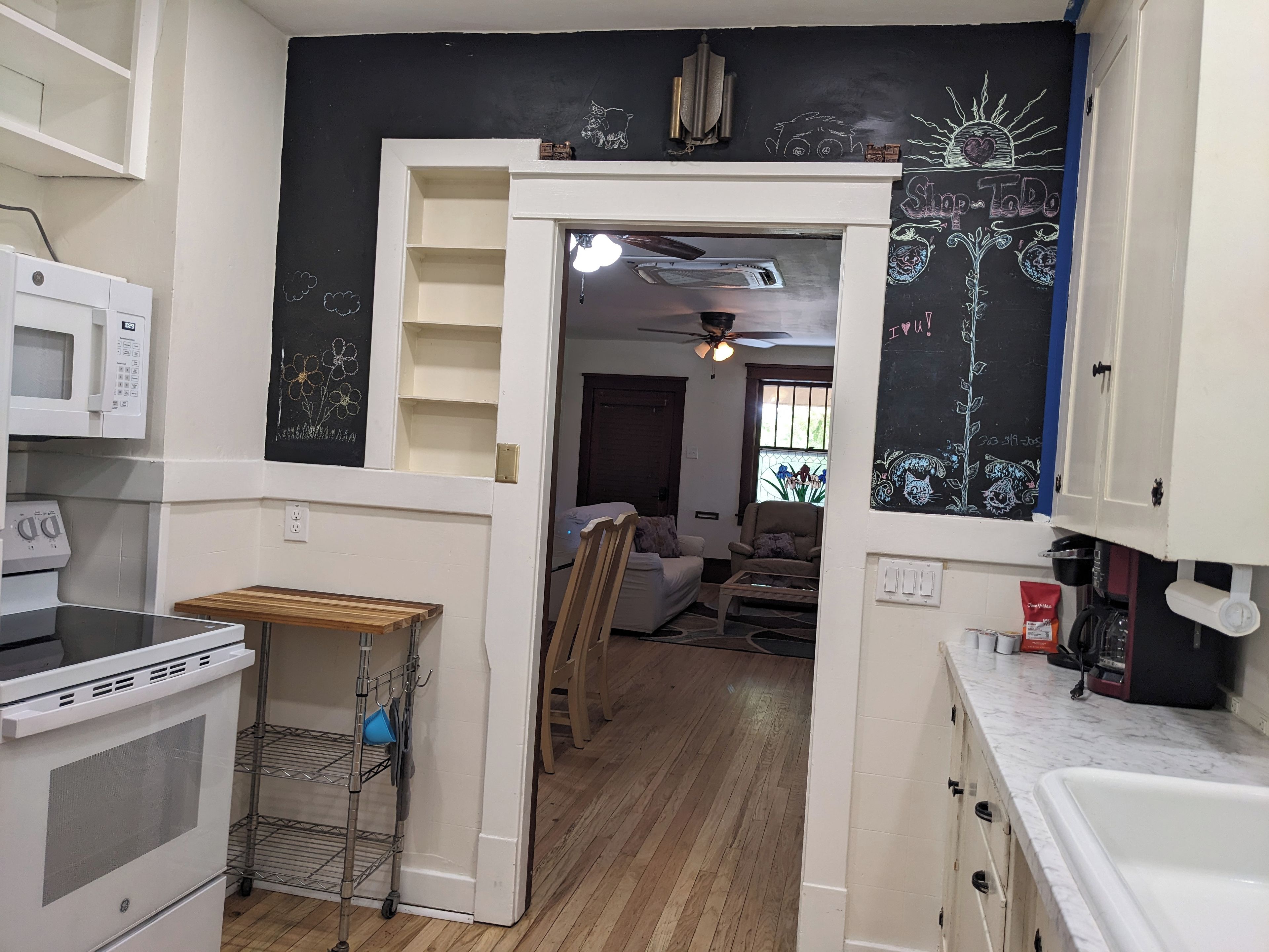 Una cocina con microondas, horno y una pequeña mesa de madera. Hay una puerta que conduce a una sala de estar con ventilador de techo y ventana.