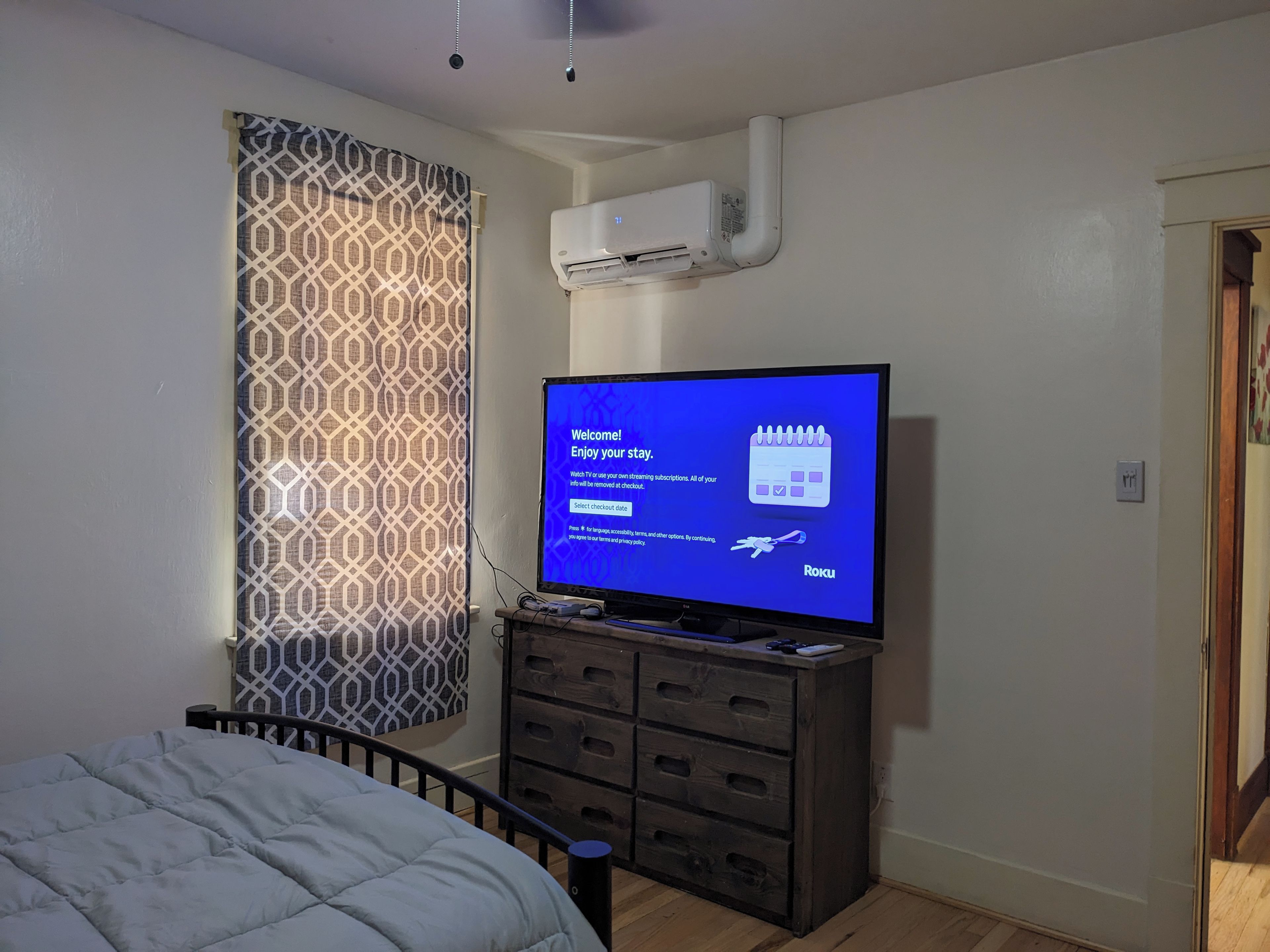 Een hotelkamer met een bed, een televisie waarop een welkomstboodschap staat, een gordijn met een patroon en een airconditioning.