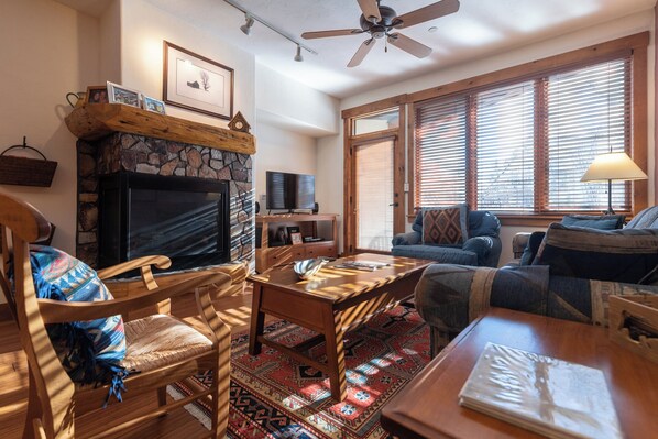 A cozy living room with a stone fireplace, wooden furniture, and large windows with blinds. - TL2102 | Warm and Cozy Home | Close to Skiing | 10 Hot Tubs | Winter Shuttle (Steamboat Springs)