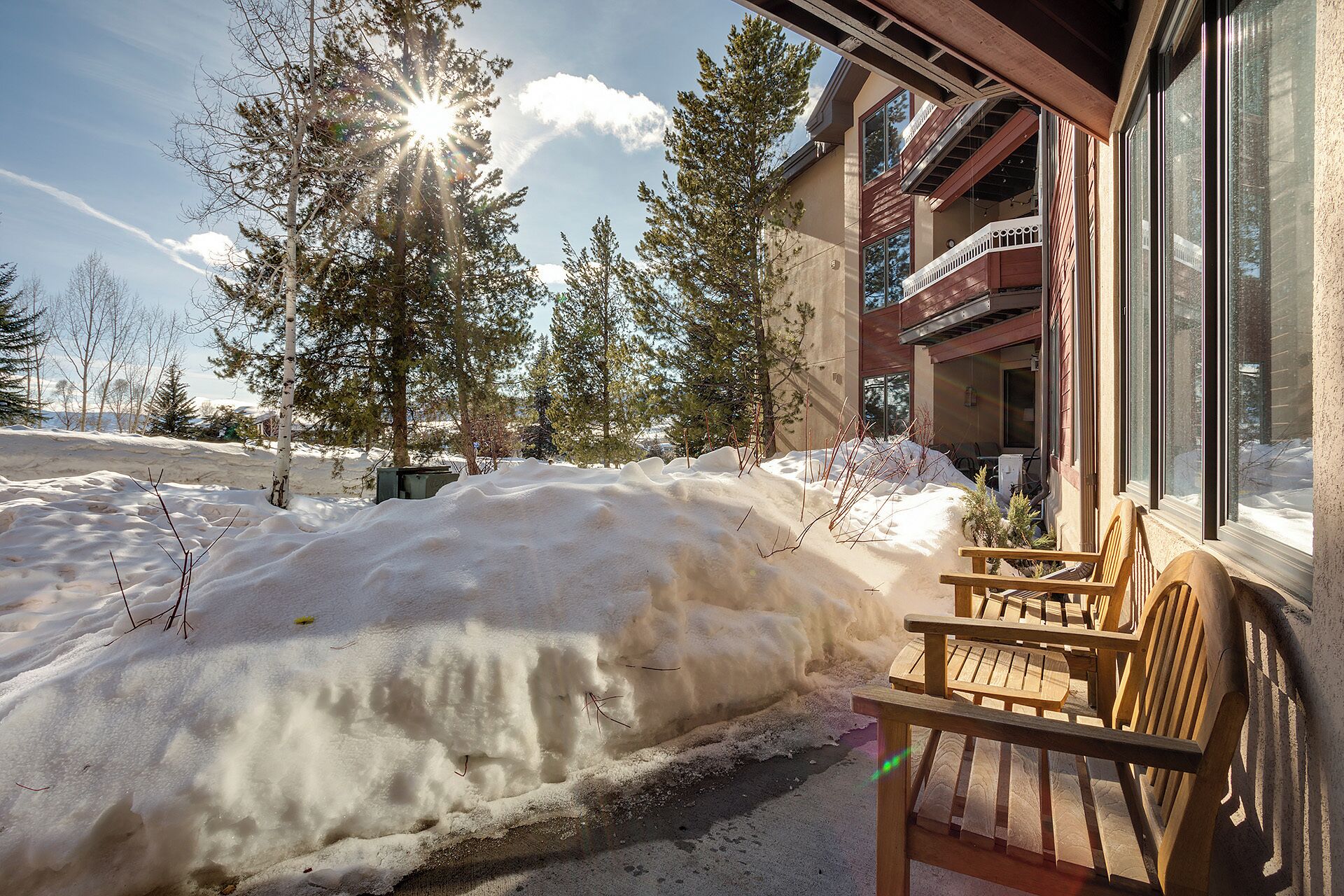 Un banco de madera frente a un edificio, con suelo cubierto de nieve y árboles al fondo.
