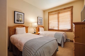A hotel room with two beds, a wooden headboard, a nightstand with a lamp, a window with blinds, and a wooden dresser. - TL2103 | Easy Access to Slopes | Beautiful Décor | Winter Shuttle | Hot Tubs (Steamboat Springs)