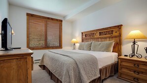 Une chambre à coucher avec une tête de lit en bois, deux lampes de chevet, un téléviseur posé sur une commode en bois et une fenêtre avec des stores.