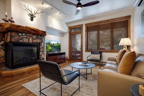 A living room with a stone fireplace, a television, a ceiling fan, and a large window with blinds. - TL2205 | Beautifully Appointed Vacation Home | Close to Skiing | A/C (Steamboat Springs)
