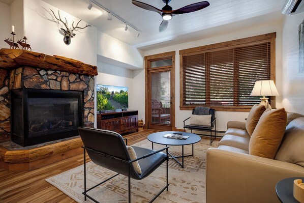 A living room with a stone fireplace, a television, a ceiling fan, and a large window with blinds. - TL2205 | Beautifully Appointed Vacation Home | Close to Skiing | A/C (Steamboat Springs)