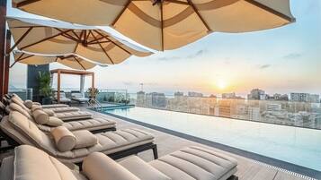 A rooftop pool area with lounge chairs and umbrellas, overlooking a cityscape at sunset.