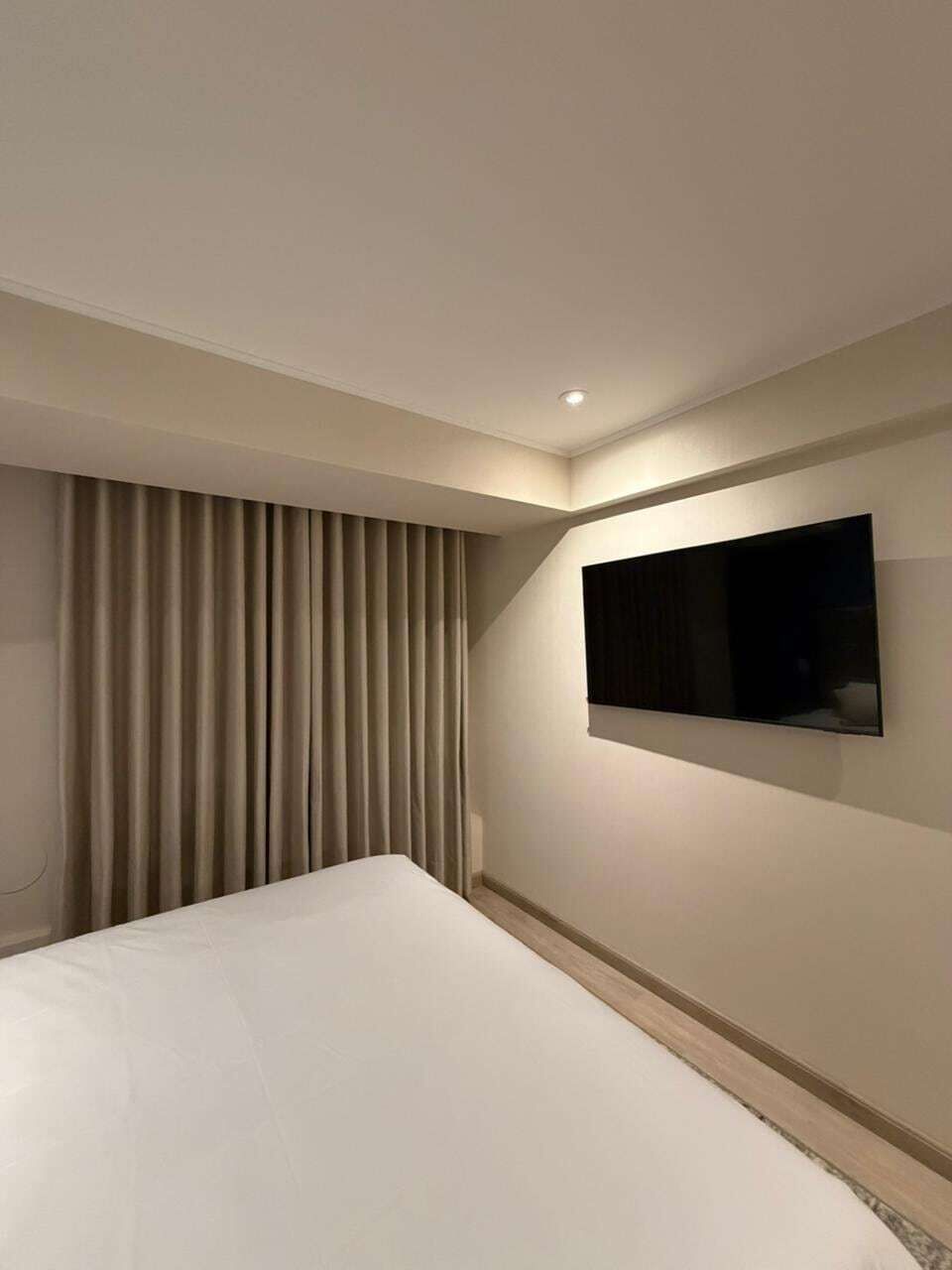 A hotel room with a bed, a wall-mounted TV, and a window with curtains.