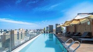Ein Infinity-Pool auf dem Dach mit Blick auf die Stadt, Liegestühle und Sonnenschirme.