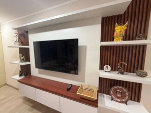 Ein modernes Wohnzimmer mit einem Flachbildfernseher an einer weißen Wand, Holzregalen mit Dekoration und einem Holztischchen mit Fernbedienung.