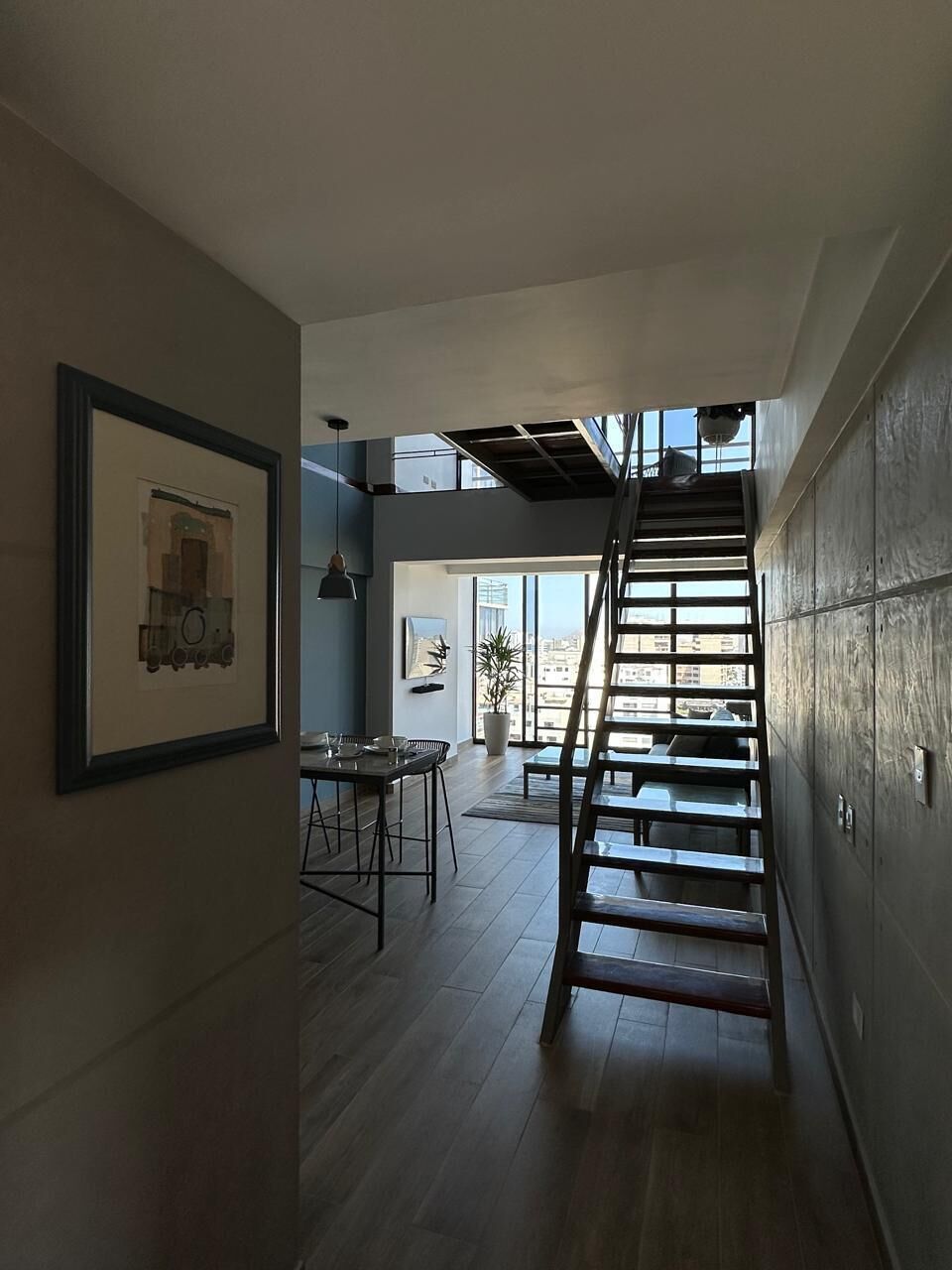 A modern interior with a wooden staircase leading to a balcony, a dining area with a table and chairs, and a framed artwork on the wall.