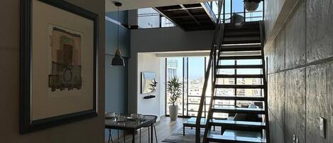 A modern interior with a wooden staircase leading to a balcony, a dining area with a table and chairs, and a framed artwork on the wall.
