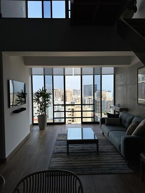 A modern living room with a large window, a sofa, a coffee table, and a potted plant. - Elegant Duplex with Terrace Sea View (Miraflores)