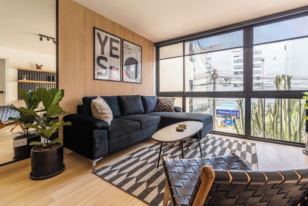 Ein modernes Wohnzimmer mit einem schwarzen Sofa, einem Couchtisch und einem großen Fenster mit Blick auf Gebäude und Kakteen.