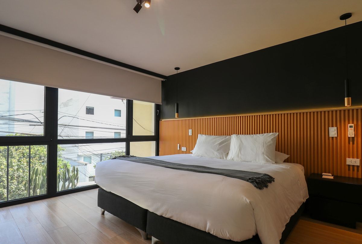 Une chambre à coucher moderne avec un grand lit, une tête de lit en bois et une vue dégagée sur l’extérieur grâce à une grande fenêtre.