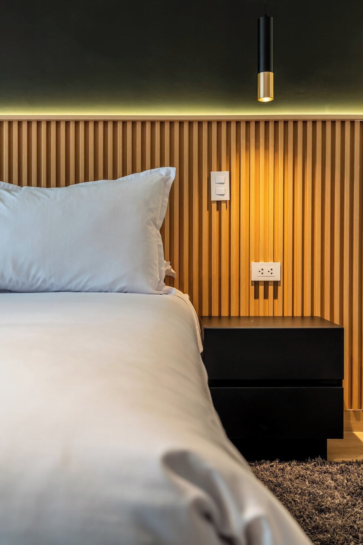 Un lit bien fait, avec une literie blanche, une table de chevet noire et un luminaire fixé au mur.