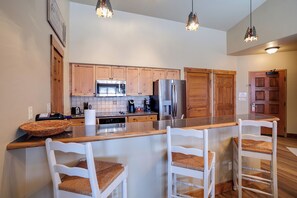 Eine Küche mit Holzausstattung, einem Edelstahl-Kühlschrank, einer Mikrowelle und einer Kaffeemaschine. Zwei weiße Stühle mit Geflocht sitzen an einer Holz Arbeitsplatte.
