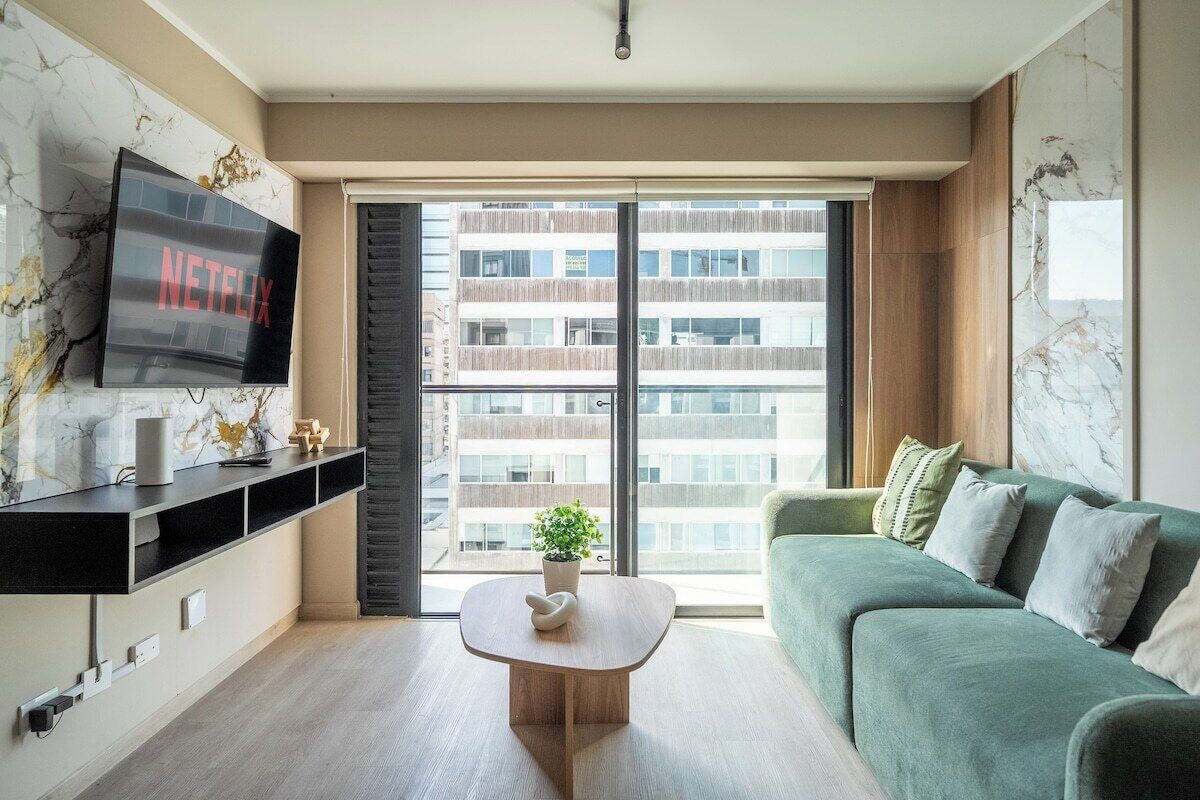 Un soggiorno moderno con una TV a schermo piatto, un divano verde, un tavolino da caffè e un'ampia finestra con vista su un edificio.