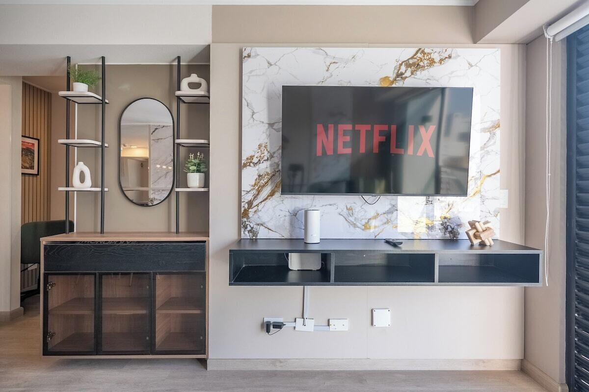 Un soggiorno moderno con TV a parete che mostra 'NETFLIX', un mobile contenitore in legno con ante in vetro, un tavolino consolle nero e una parete con decoro marmorizzato.
