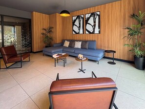 En moderne stue med sofa, to lænestole, et sofabord og et tilhørende sofabord. Der er abstrakte malerier på væggen og en potteplante.