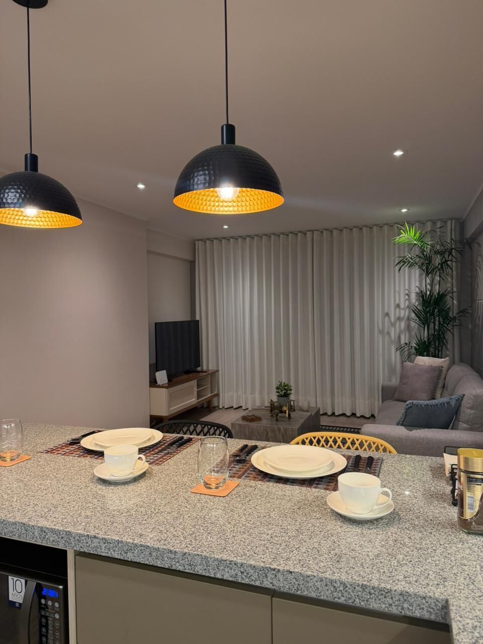 A modern kitchen with granite countertops, a dining area with a table set for two, and a living room with a sofa and curtains.