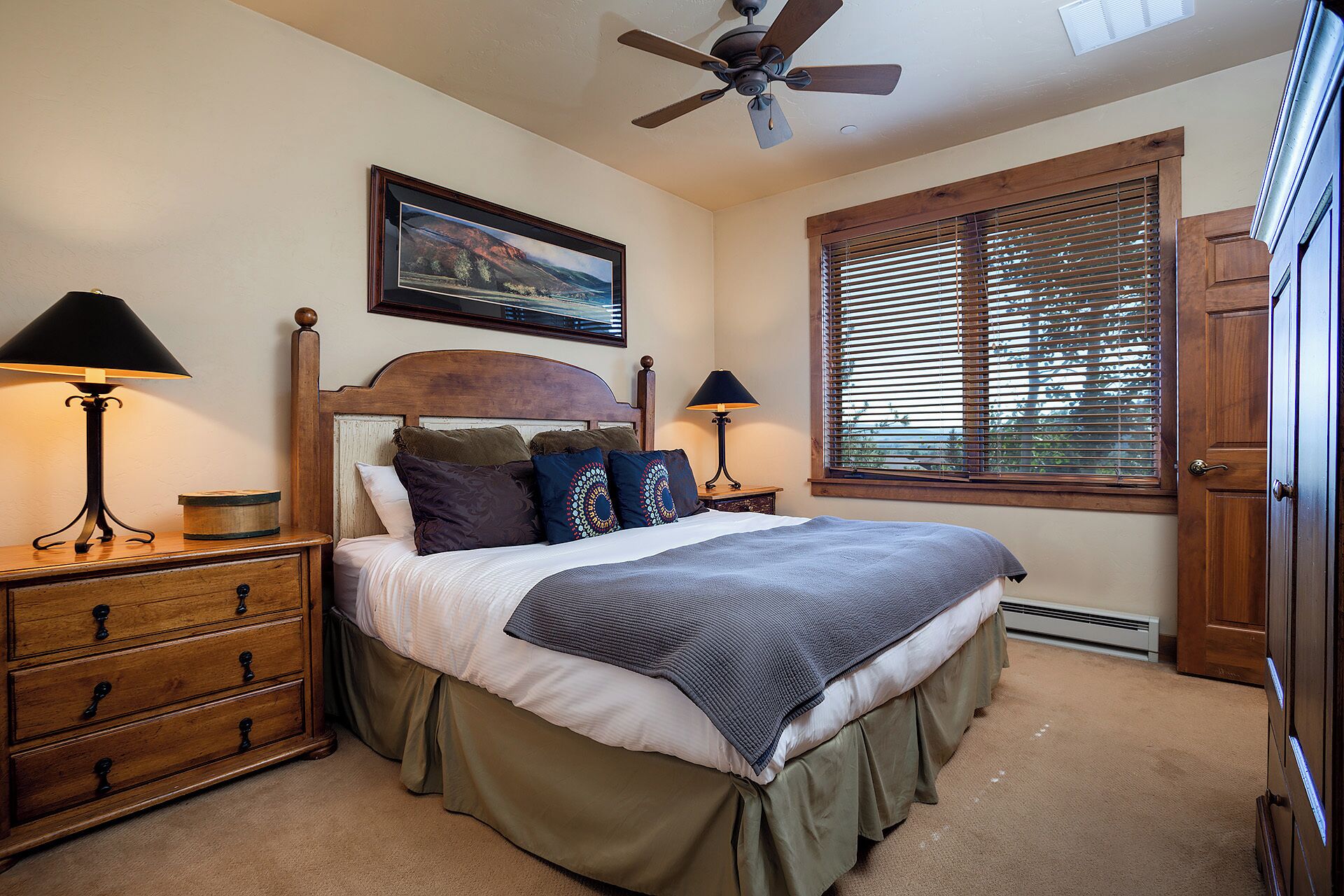A bedroom with a bed, two bedside tables, a ceiling fan, a window with blinds, and a door.