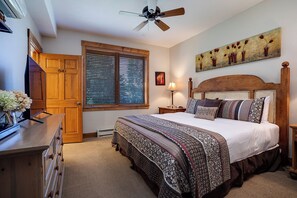 A bedroom with a bed, wooden headboard, a ceiling fan, a window with blinds, a painting on the wall, and a dresser with a TV and flowers. - TL2304 | Beautifully Appointed | Vaulted Ceilings | A/C | 10 Hot Tubs (Steamboat Springs)