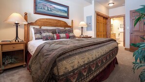 Un dormitorio con una cama grande, dos mesitas de noche, una mesita auxiliar, un cabecero de madera, un cuadro enmarcado en la pared y una puerta que da a otra habitación.