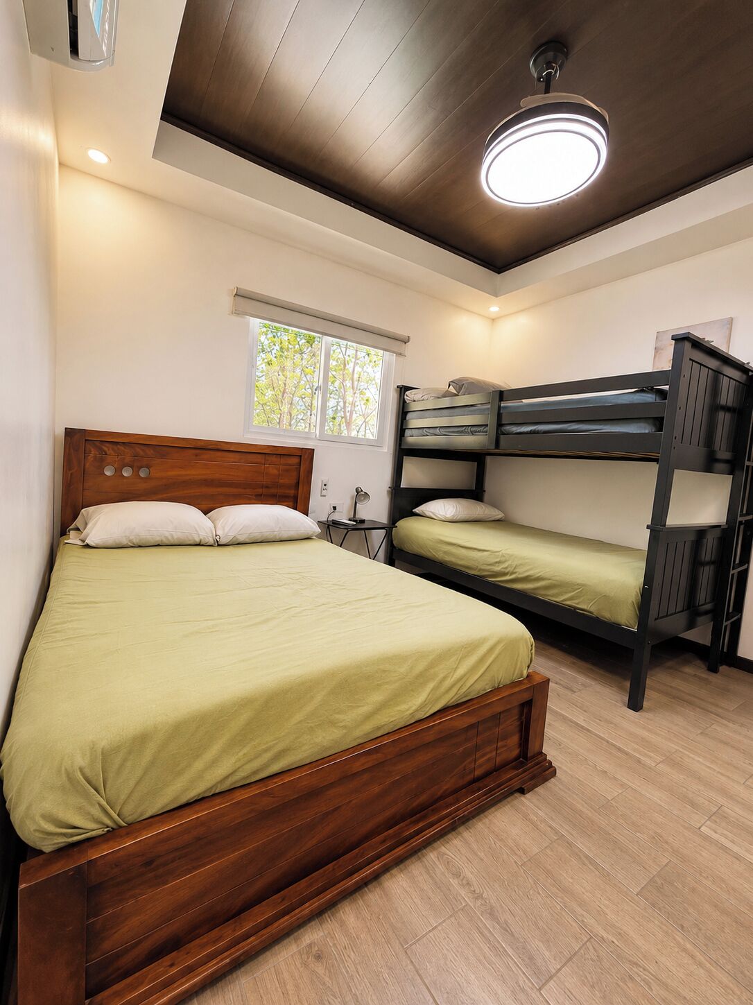 Habitación con una cama individual y una litera, piso de madera, una lámpara en el techo y una ventana con persianas.