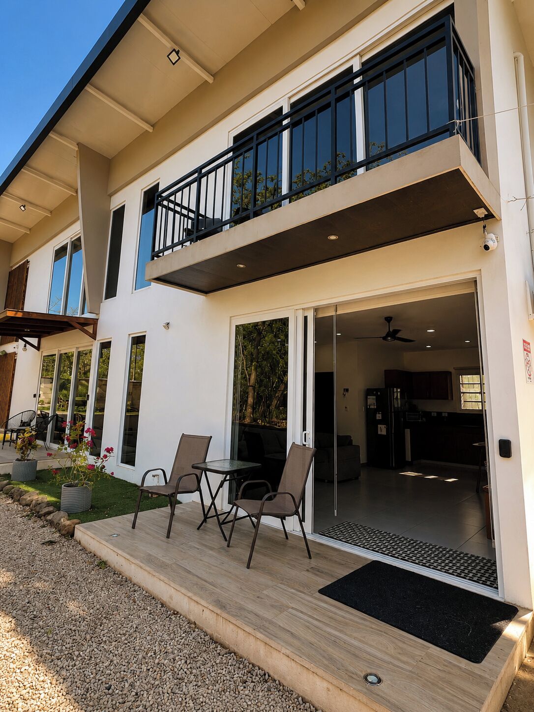 A modern two-story house with a balcony, a patio with seating, and a sliding glass door leading to an interior space.