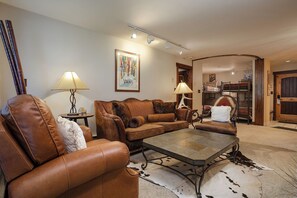 Ein Wohnzimmer mit Ledersofas, einem Couchtisch und einer Lampe. An der Wand hängt ein Gemälde, im Hintergrund steht ein Etagenbett.