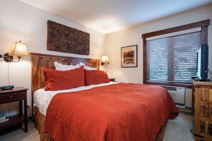 Habitación de hotel con cama, lámparas de noche, una obra de arte en la pared, ventana con persianas y televisor sobre un soporte de madera.