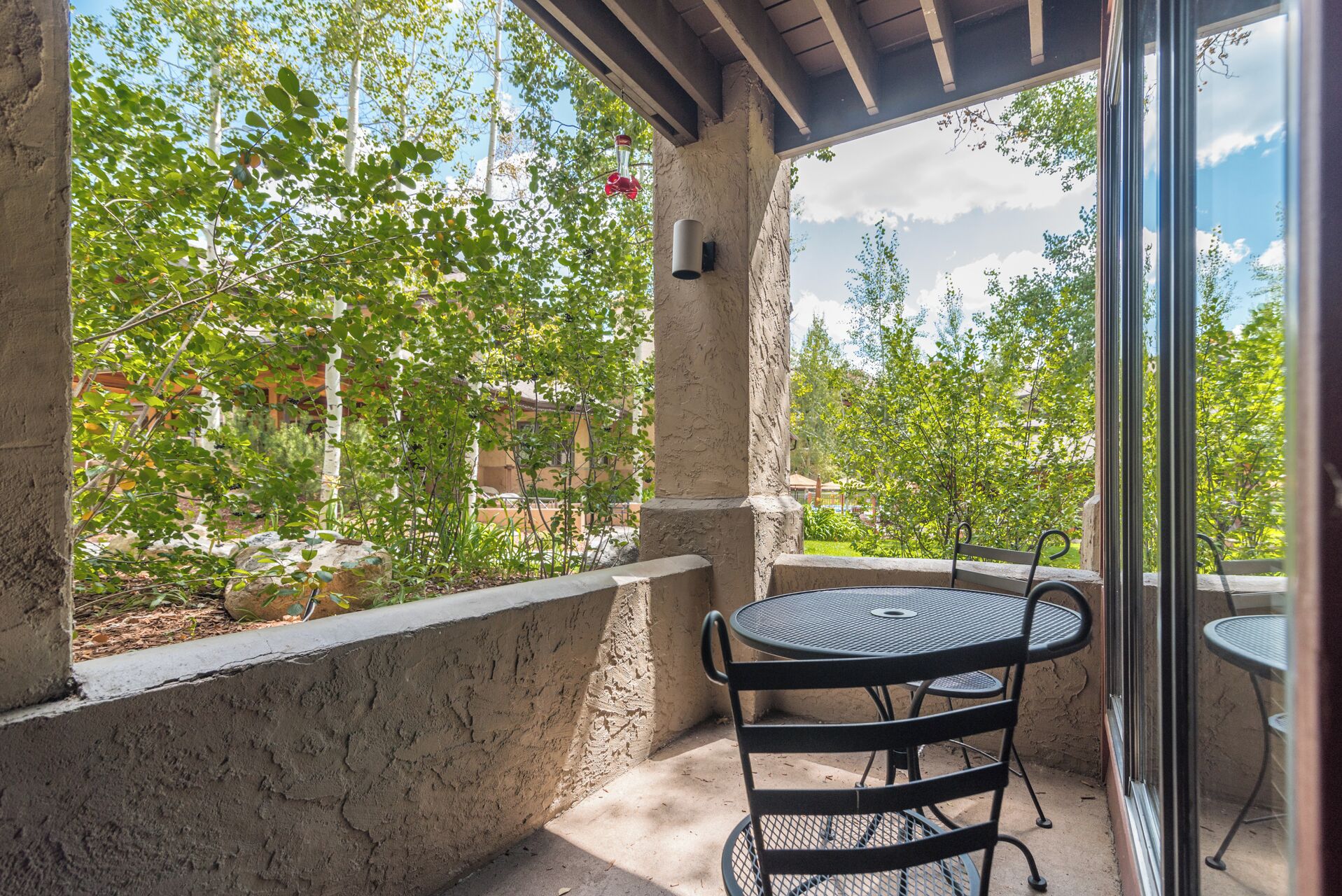 Un patio techado al aire libre con una mesa y sillas de metal redondas, rodeado por un muro de piedra y abundante vegetación.