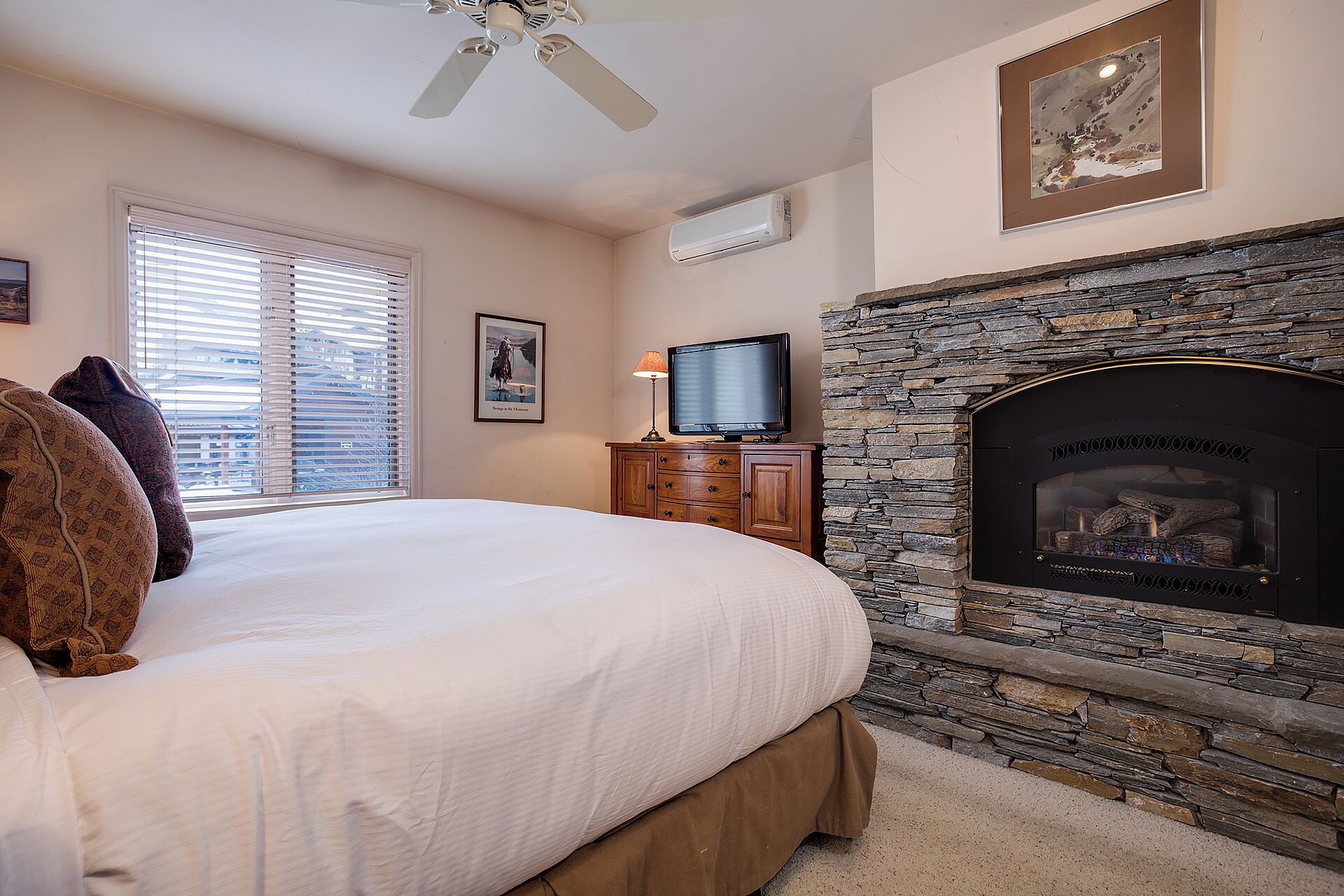 A bedroom with a bed, a stone fireplace, a television, and a ceiling fan.