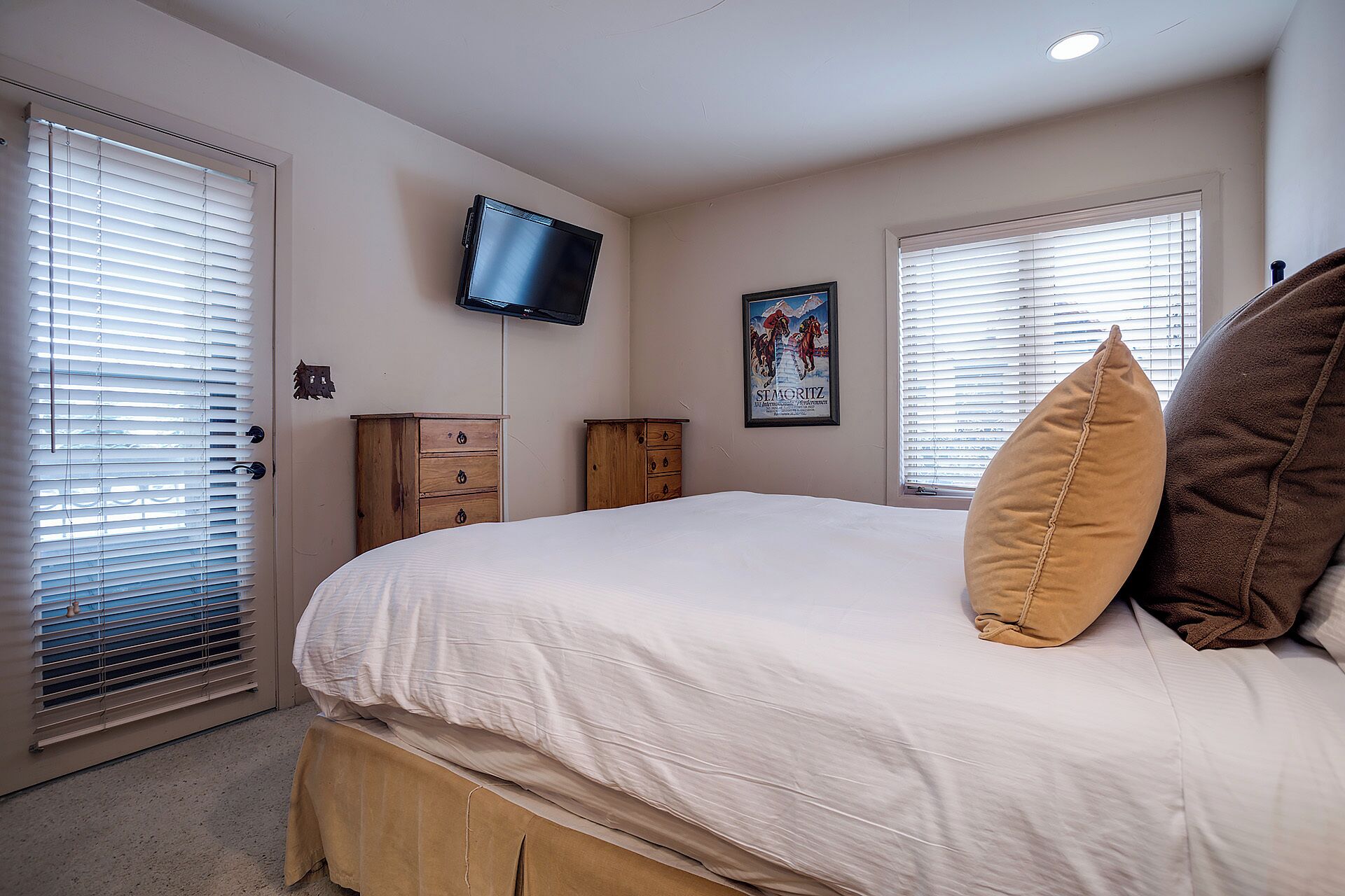 A bedroom with a bed, two wooden dressers, a television mounted on the wall, and two windows with blinds.